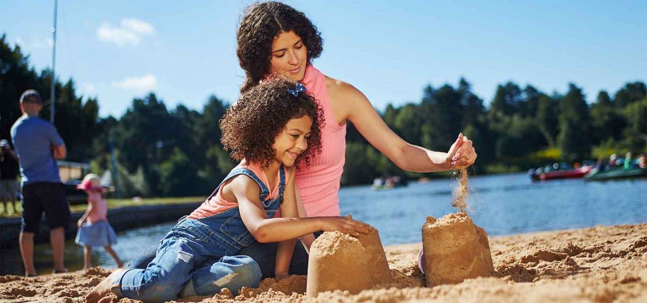 Two people build sandcastles, one pouring sand over a turret, the other shaping another mound, on a sunny lakeside beach with boats on the water and families in the background.