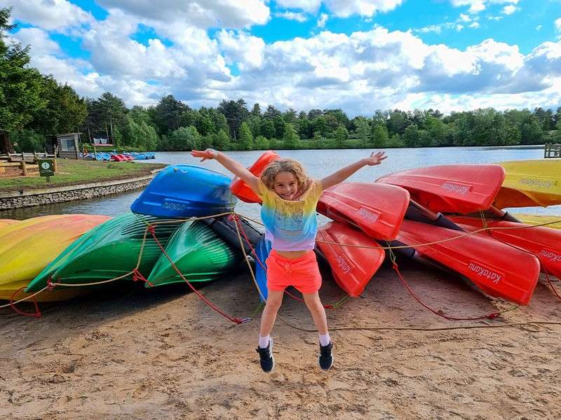 A little girl jumping on the beach in front of multicoloured canoes.