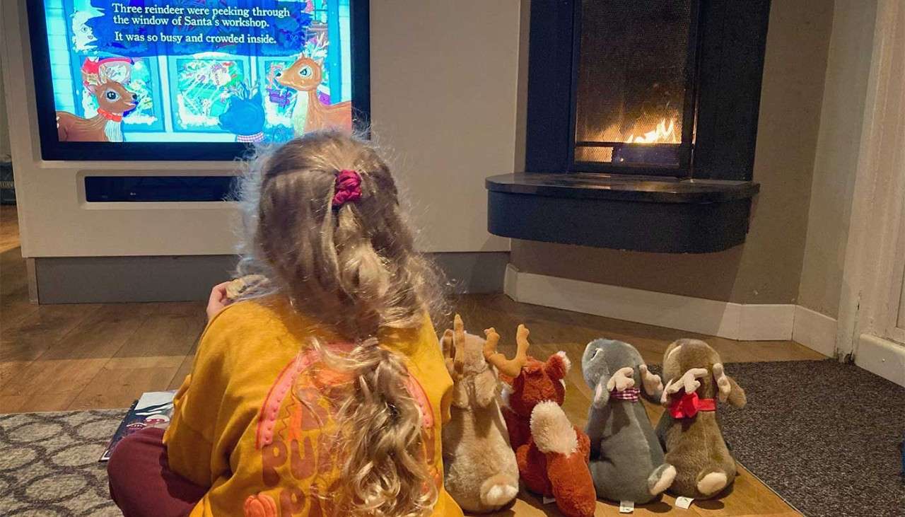 A little girl watching TV in her lodge with her Christmas teddies sat next to her.