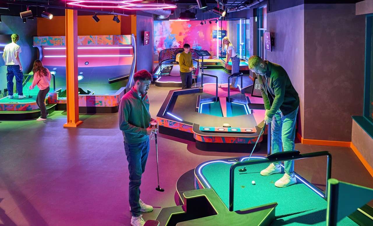 Mini-golf players putt balls across neon-lit, futuristic courses; one man prepares a shot while others wait and chat, inside a colorful indoor entertainment venue with glowing obstacles and geometric platforms.