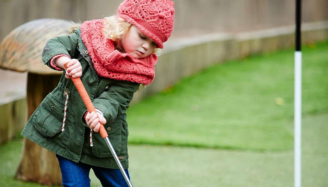 Child holds a putter and swings gently, concentrating on a mini-golf green. Bundled in a pink knit hat and scarf with a green coat, near a black-and-white flagstick outdoors.