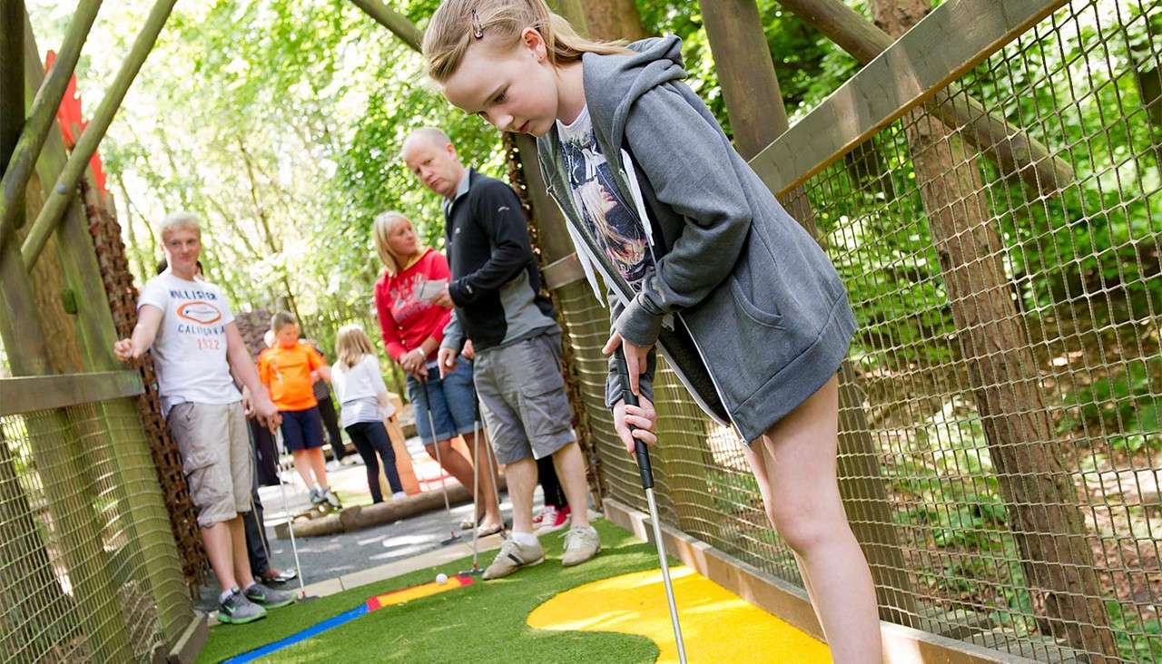 Girl putts a mini-golf ball on an outdoor course, surrounded by wooden fencing and trees, while several people wait behind her. Text: HOLLISTER CALIFORNIA 1922.