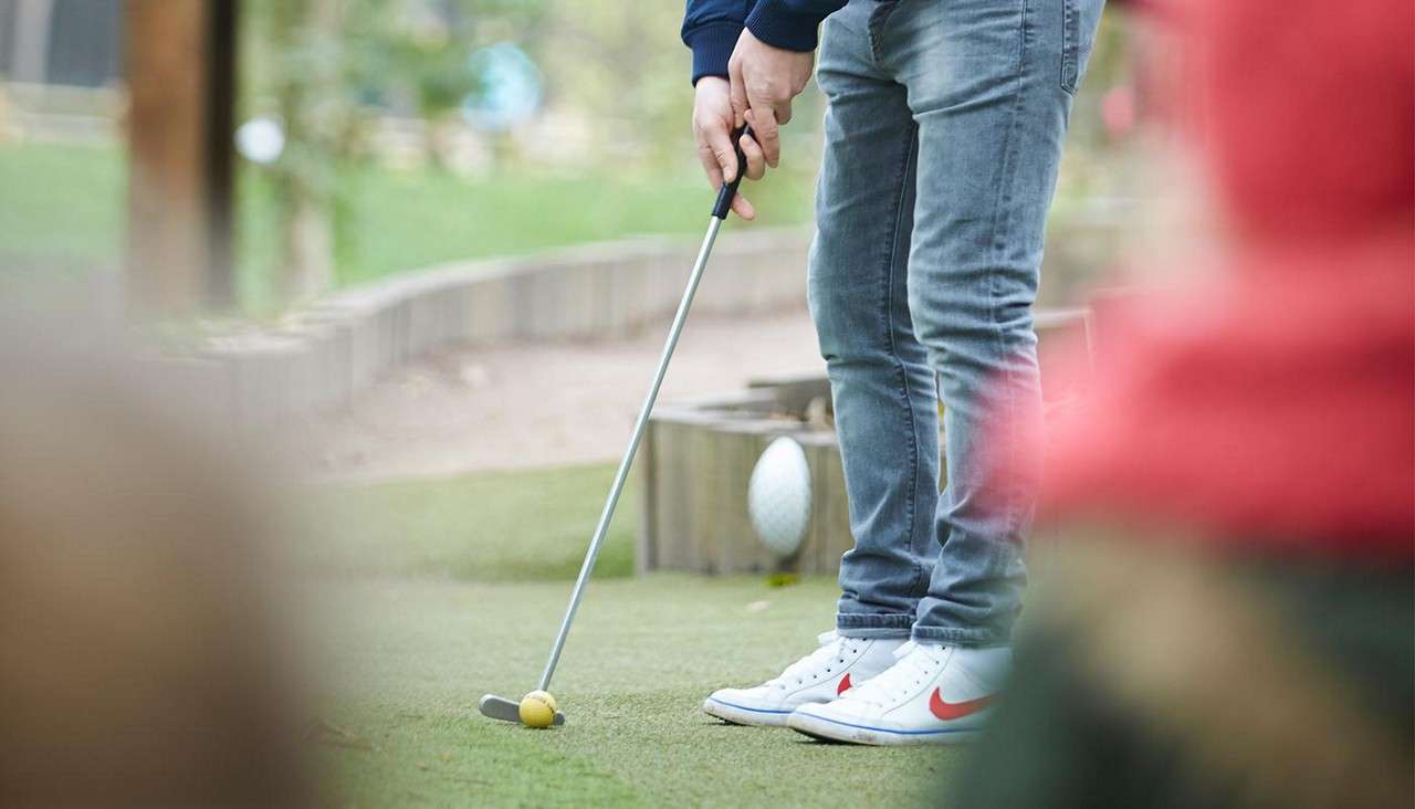 Person holds a putter, addressing a yellow ball on artificial turf; wearing jeans and white sneakers. Scene occurs at an outdoor mini-golf course bordered by wooden edging, with blurred foreground onlookers.