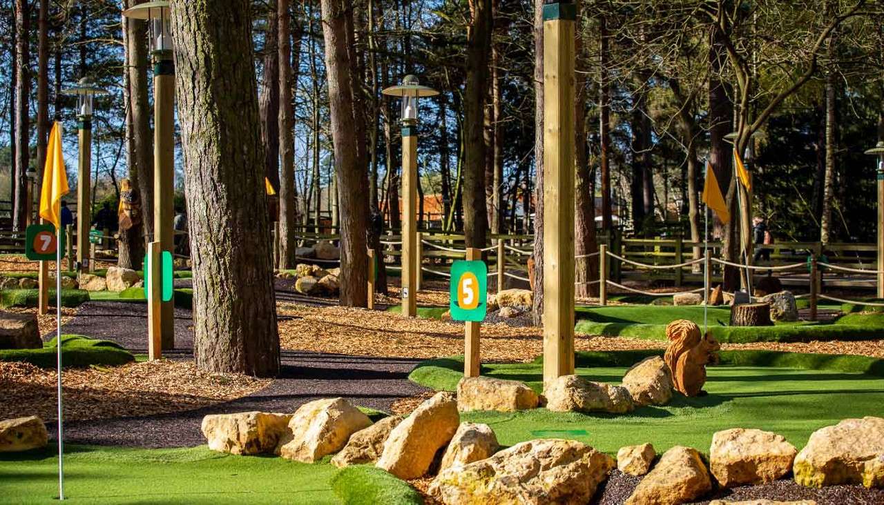 Mini-golf holes display flags and numbered markers, guiding play. In a wooded park, artificial turf winds around rocks, rope-fenced paths, and lamp posts under tall trees. Visible text: 5, 7.