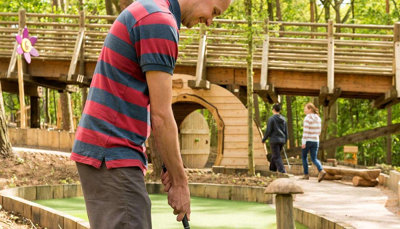 Man with a putter lines up a shot, playing mini-golf on a green lane. Around him, two people walk near a wooden bridge and tunnel in a forested course. Sign reads "6".
