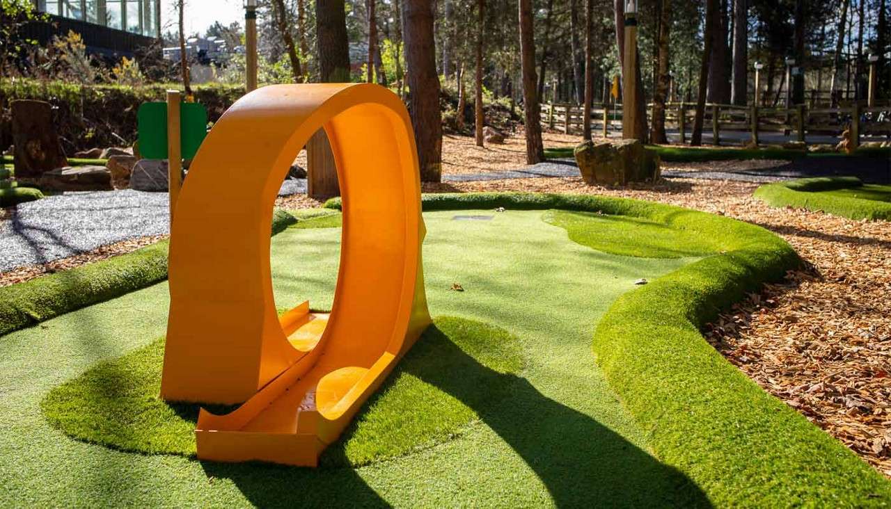 Bright orange loop obstacle sits on a mini-golf hole, channeling balls through its circular tunnel; surrounding context is an outdoor course with artificial turf, woodchip paths, trees, and sunlight.