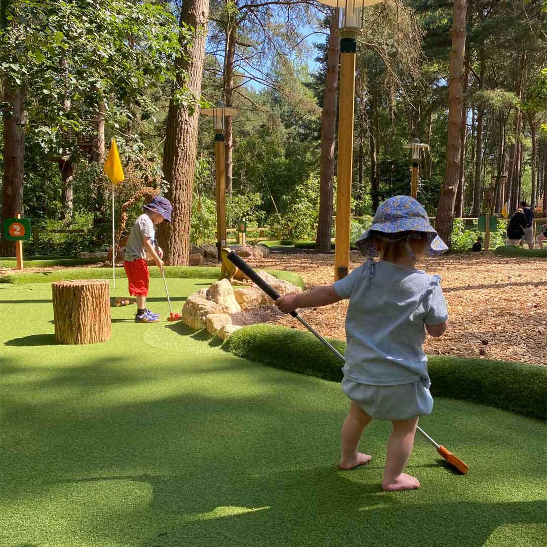 Toddler holds a mini-golf putter, walking barefoot toward a green. Another child putts near rocks and a yellow flag. The course sits in a wooded park with sign: “2”.