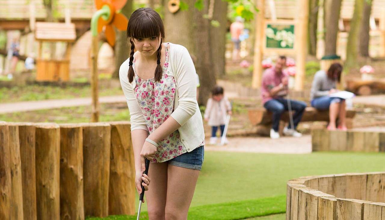 Teen girl with pigtails swings a putter, aiming at a mini-golf hole; wooden barriers and artificial turf surround her, with blurred park visitors and trees in the background.