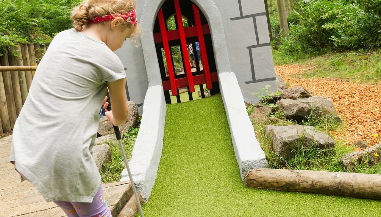 Child golfer putts toward a castle-themed mini-golf tunnel with a red gate, guiding the ball up a green ramp; surroundings include wooden rails, rocks, plants, and a forest path.