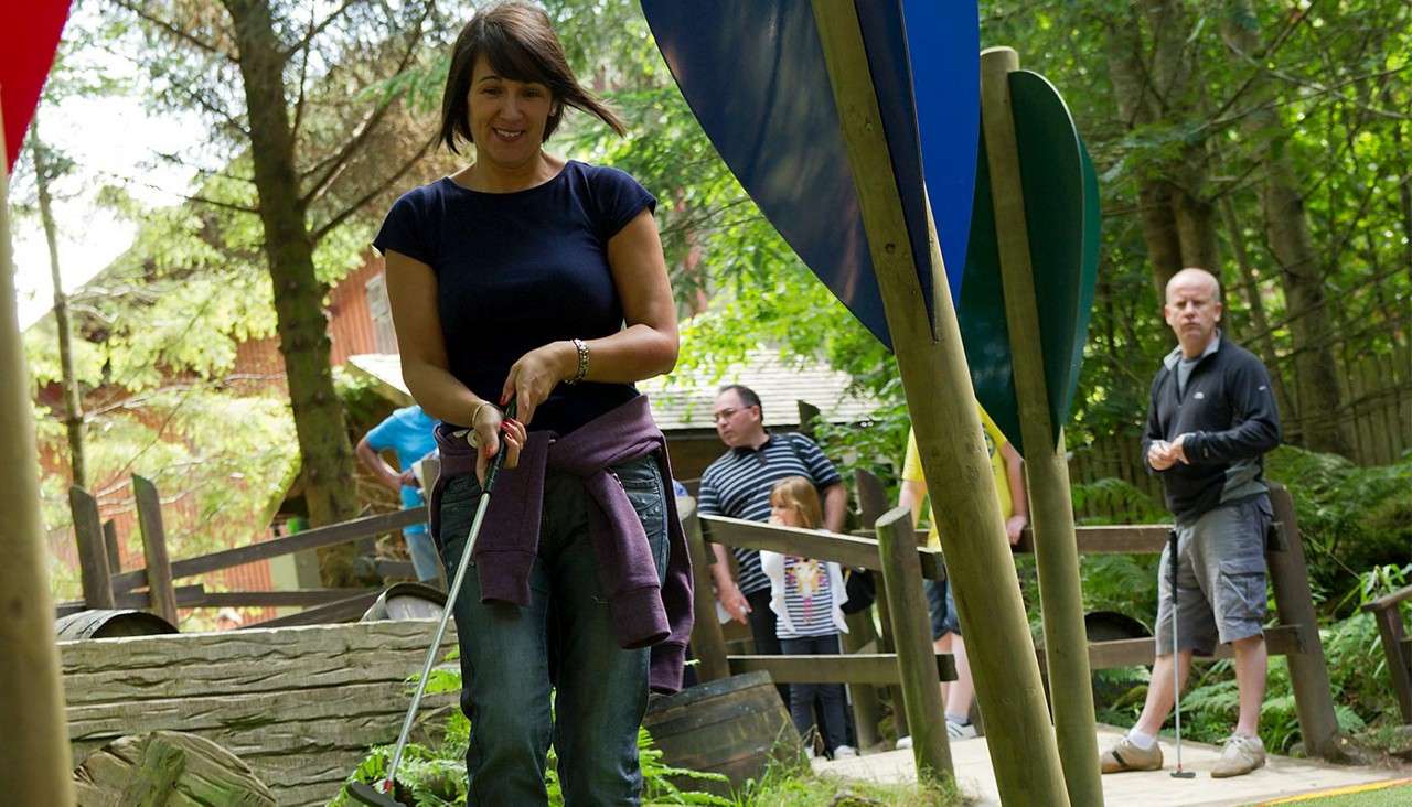 Woman swings a putter, putting a ball on a mini-golf hole; bystanders wait and watch in the background; course winds through trees, wooden fences, and barrels in a forested park.