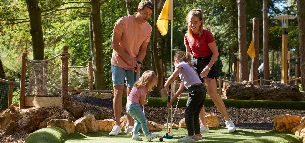 Two children putt a mini-golf ball while two adults guide them, on an outdoor course with yellow flags, artificial turf, rocks, and trees in warm daylight.