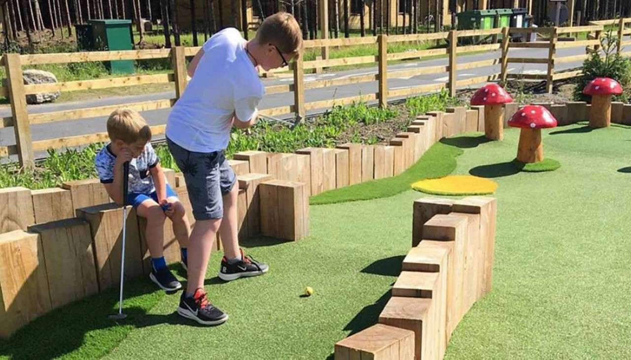 Boy putting a yellow mini-golf ball; another boy sits with a club watching, on an outdoor mini-golf course with artificial turf, wooden barriers, and red mushroom obstacles beside a fence.