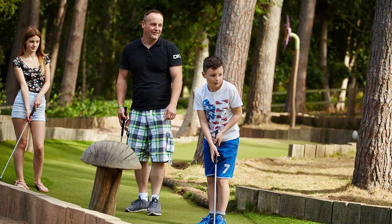 Boy putts a ball on a mini-golf hole, watched by a man as a girl lines up nearby; wooden borders, trees, and a mushroom obstacle surround them. Text: DK, 7.