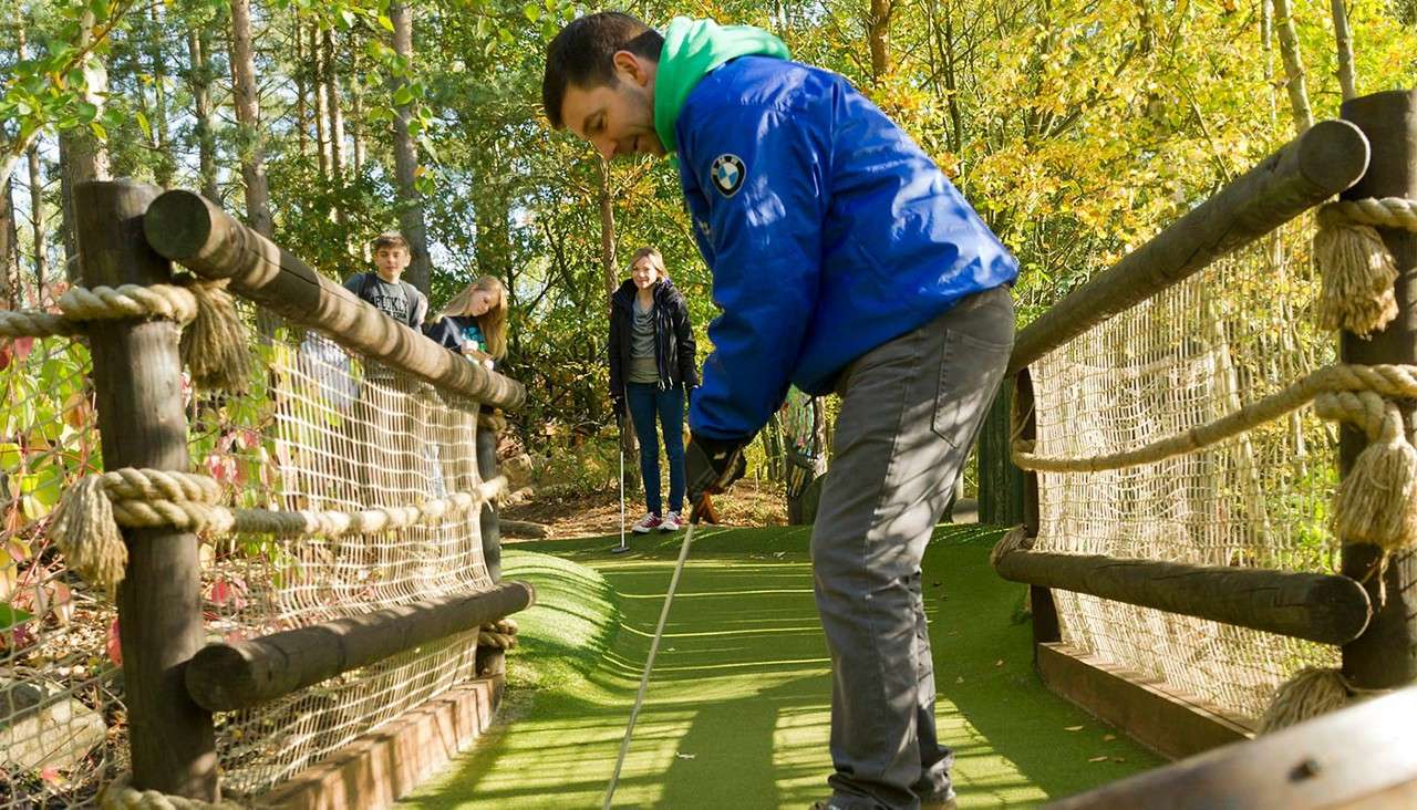 Golfer putts on a narrow, undulating mini-golf bridge, watched by three people; rope-and-net railings line the path amid sunlit trees. Text: BMW.