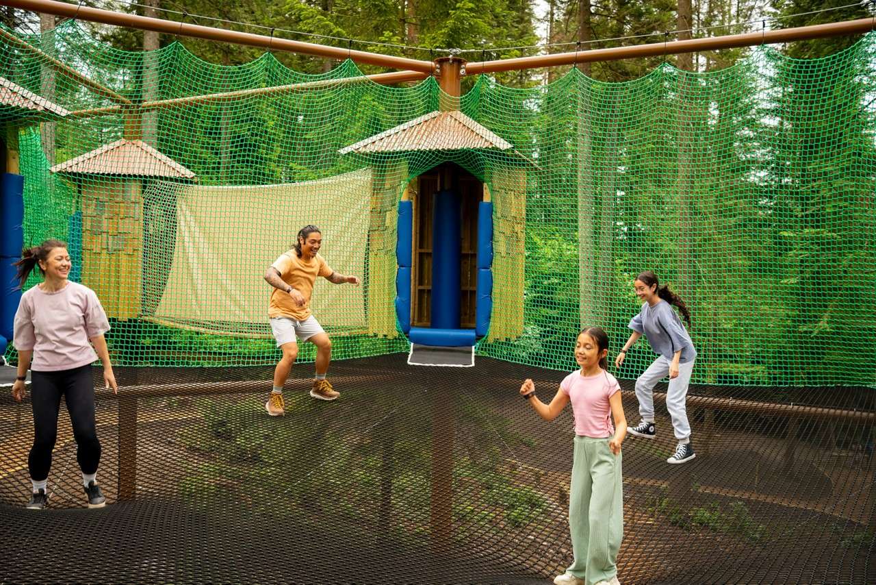 Four people bounce and walk on a giant net trampoline, surrounded by green safety nets and a small treehouse slide, high among forest trees.