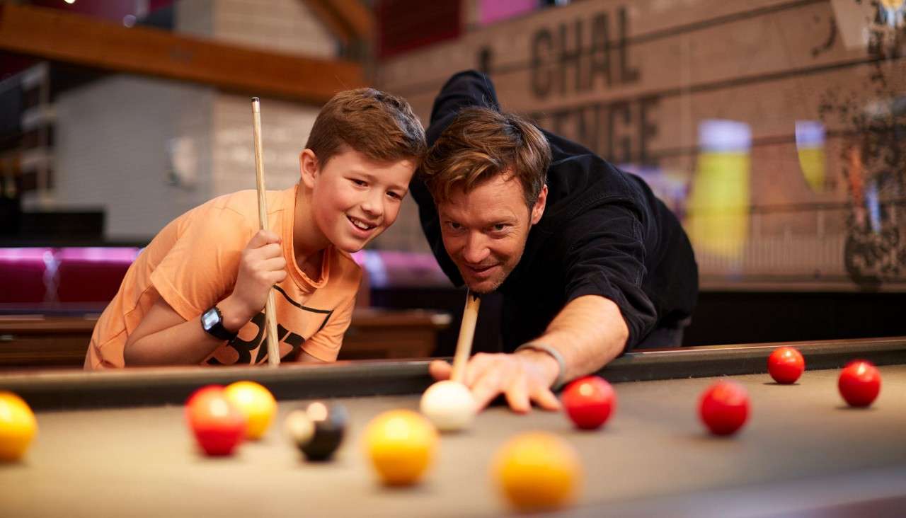Man with cue aims at white ball; boy watches closely. In a pool hall with scattered red and yellow balls. Background wall text: “CHAL LENGE”.