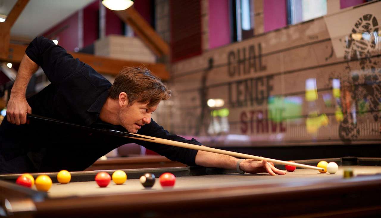 Pool player leans and aims a cue to strike a white ball; in a bar-style hall, colored balls dot the table, mural text reads: CHAL LENGE STRIVE.