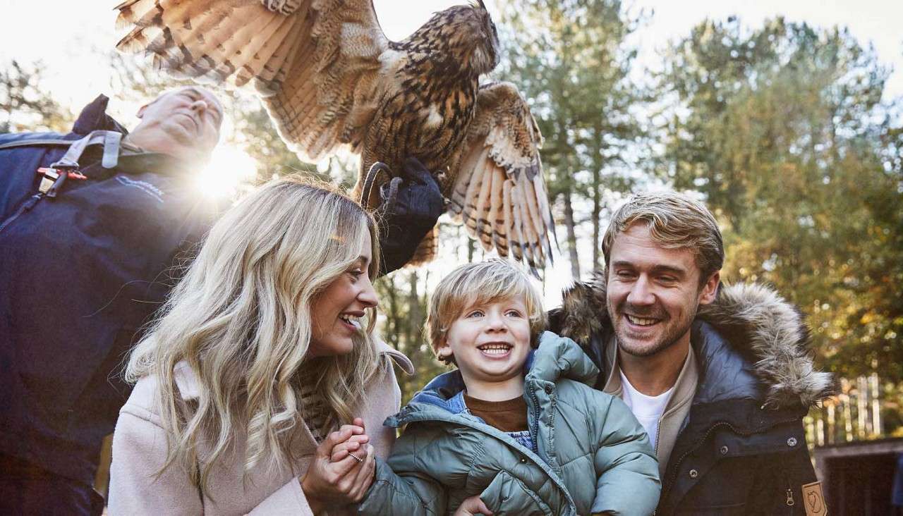 Owl spreads wings while perching on a handler’s gloved hand; a smiling child and two adults watch closely, bundled in coats, standing together in a sunlit forest setting.