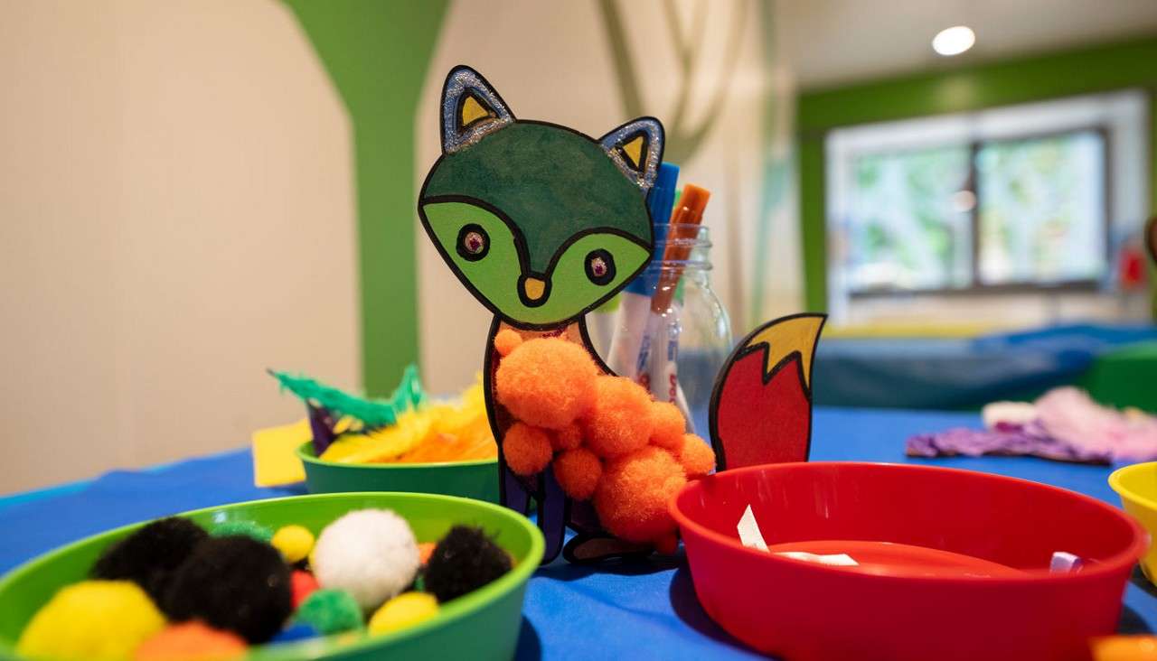 Paper fox craft shows glued orange pom-poms, standing beside a jar of brushes on a blue table, surrounded by bowls of colorful pom-poms and feathers in a bright, child-friendly classroom.