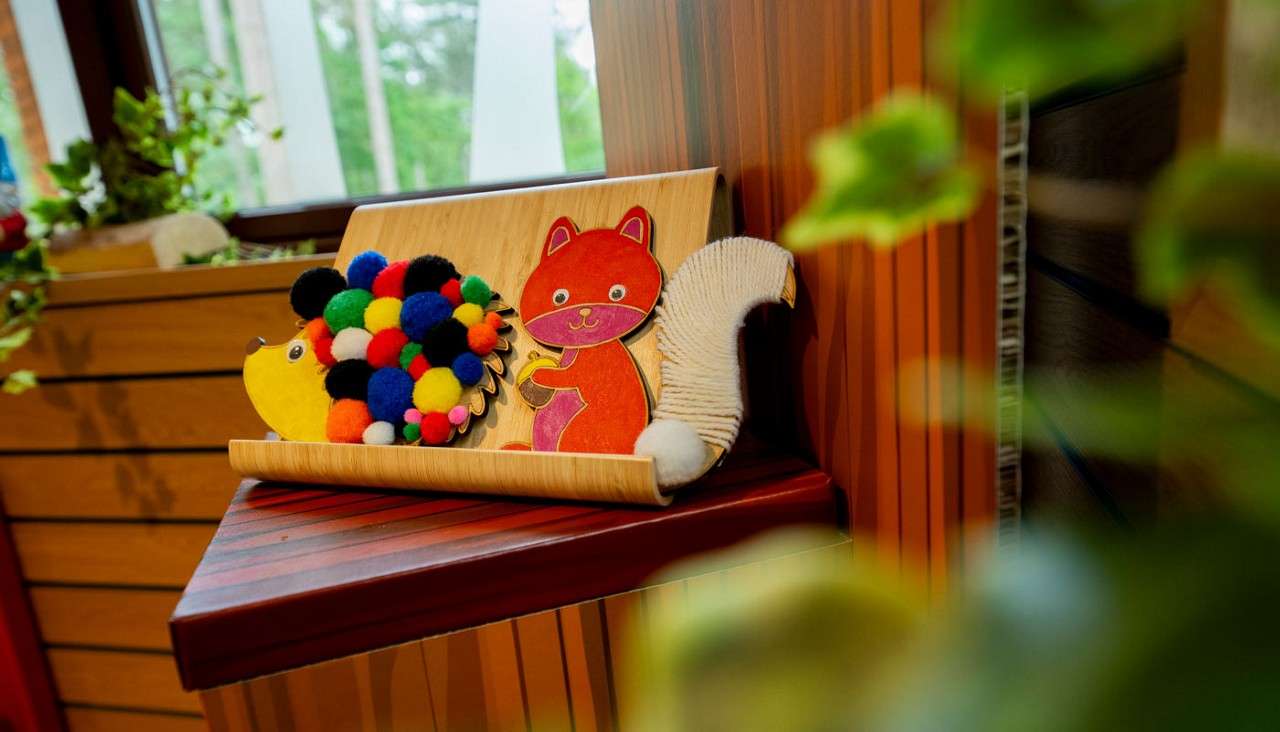 Colorful children's craft animals sit propped on a small wooden shelf: a pom-pom-covered hedgehog and a cartoon squirrel with a bristled tail, displayed in a plant-filled room near a window.