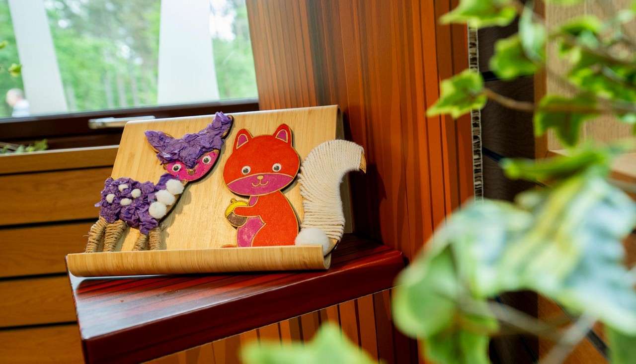 Two handmade animal cutouts displayed on a wooden stand: a purple deer and an orange squirrel holding an acorn, on a corner shelf near a window with green leaves foreground.
