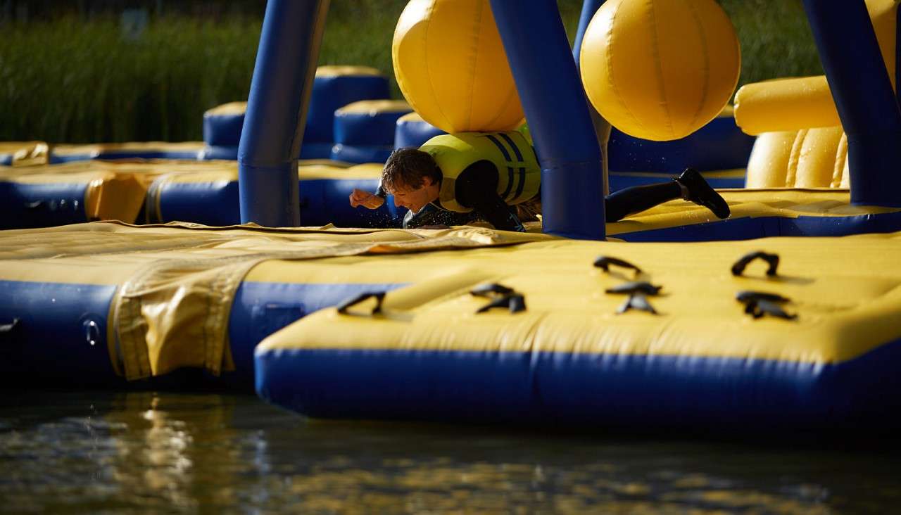 Person wearing a life jacket crawls across a yellow-and-blue inflatable water obstacle course, dodging large hanging balls, above shallow water with reeds in the background.