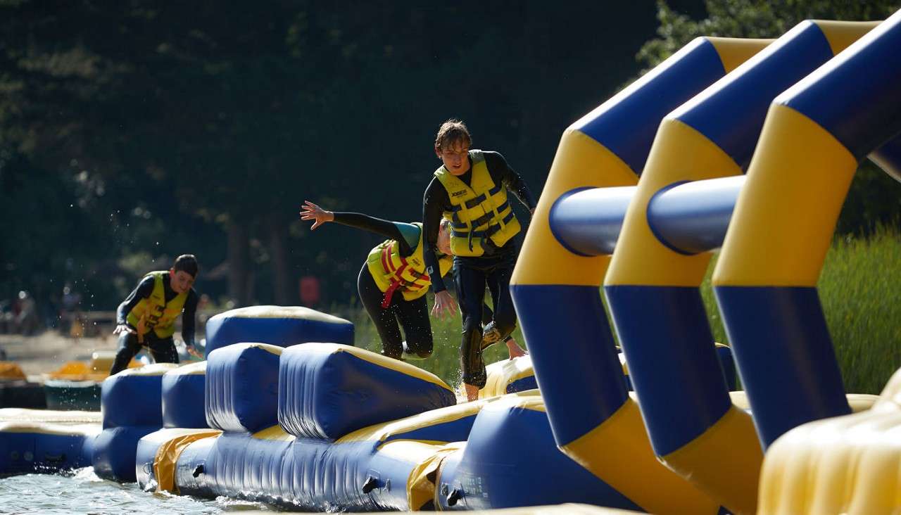 People in yellow life vests run and balance on a blue-and-yellow inflatable obstacle course, splashing as they cross floating platforms on a lake, with trees and reeds in the background.