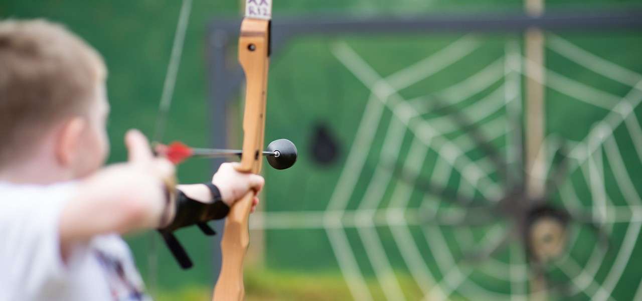 Bow aims a suction-cup arrow; action: a person draws to shoot; context: outdoor archery range with green backdrop and spiderweb target. Text visible: K12.