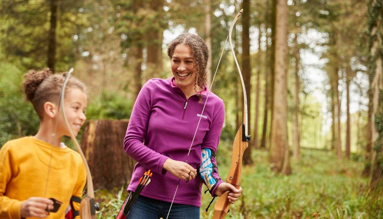 Two archers hold recurve bows and chat, smiling, with arrows in a hip quiver. They stand among tall trees on a sunlit forest trail, suggesting a casual outdoor archery session.