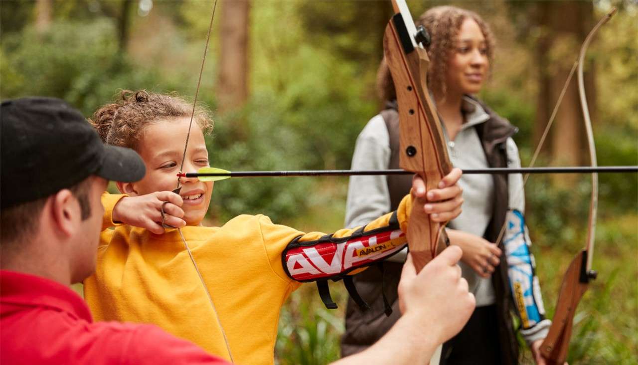 Child archer draws a bow, guided by an instructor, while aiming an arrow; another observer stands nearby in a wooded outdoor setting. Text: AVALON.