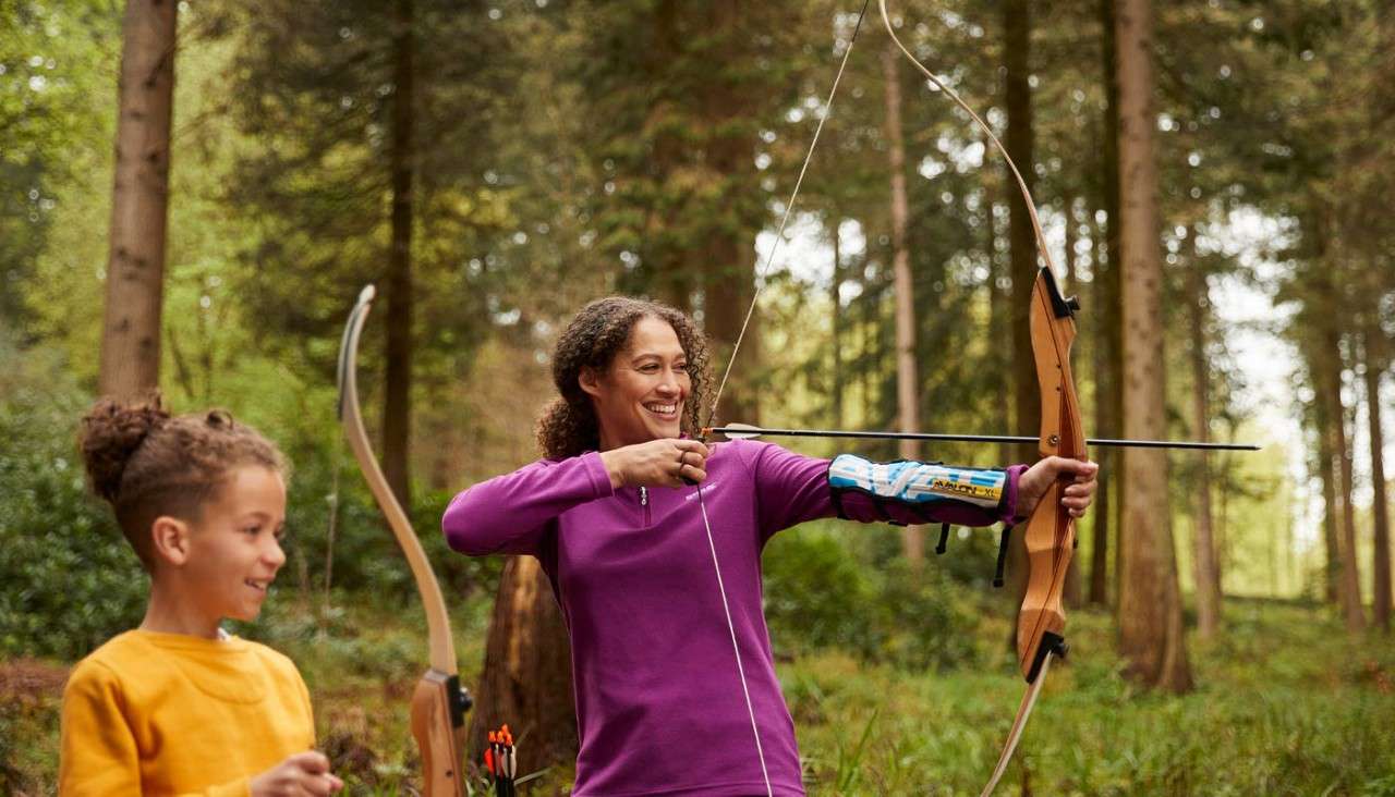 Archer draws a recurve bow, smiling while nocking an arrow; a child watches holding another bow. They stand in a sunlit forest. Visible text on armguard: AVALON.
