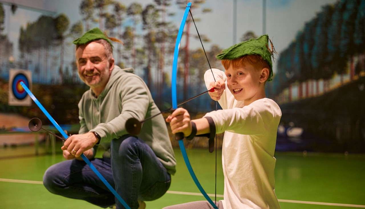Child archer aims a blue bow with suction-cup arrow, smiling, while an adult beside them crouches with a bow; both wear green hats in an indoor range with target backdrop.
