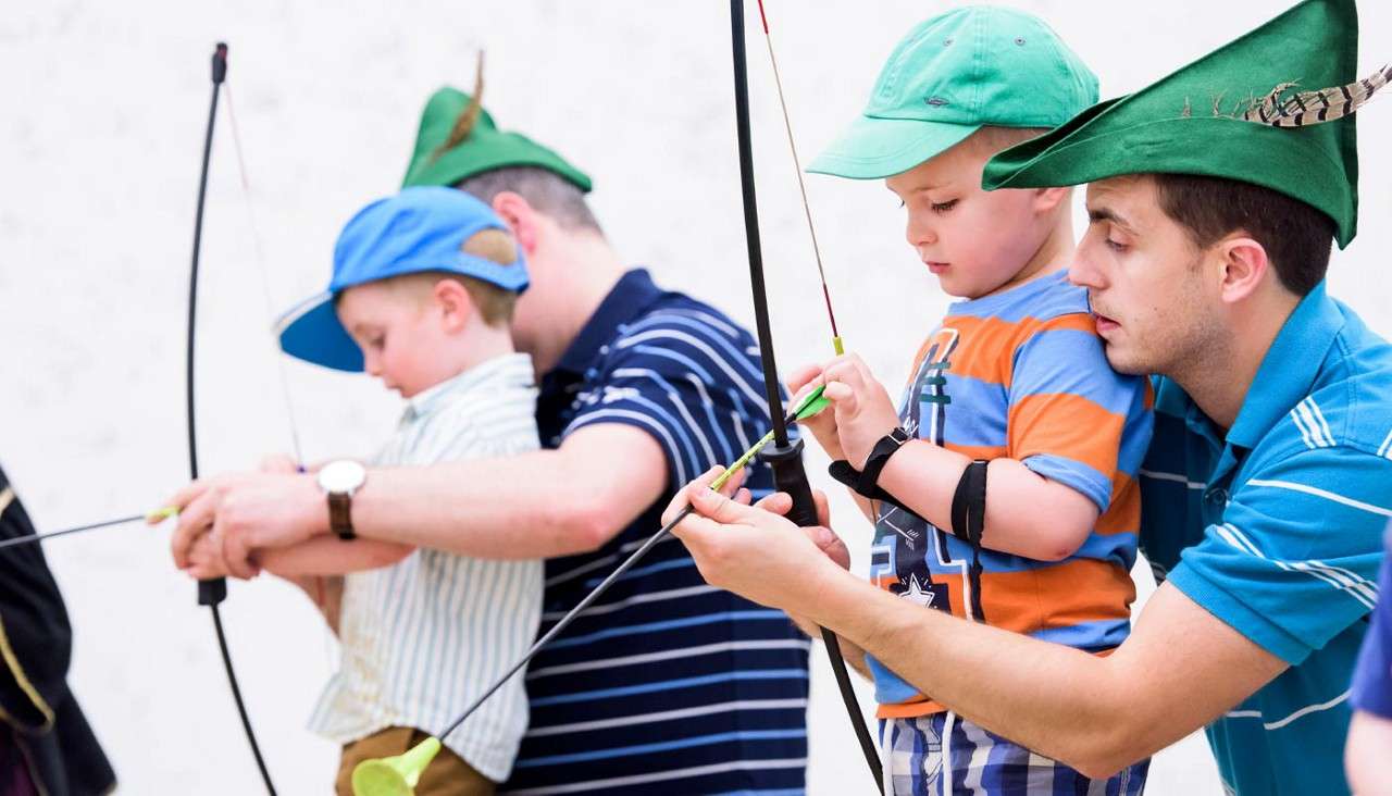 Children draw bows while adults guide their hands, practicing archery indoors. One child wears a green cap; another adult wears a feathered green hat. Bows are nocked with suction-cup arrows.