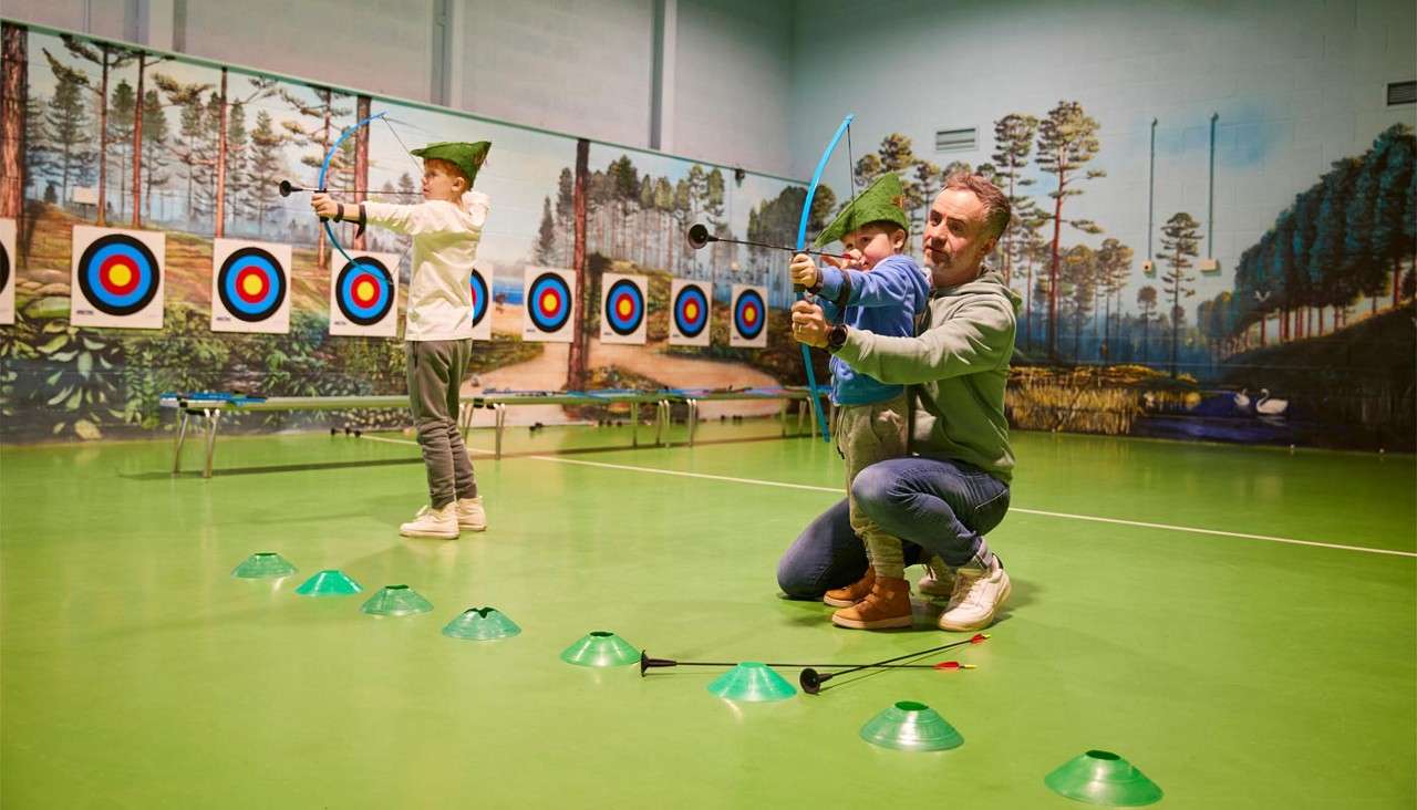 Two children with toy bows shoot suction-cup arrows while an adult guides one kid; archery targets line a gym with a forest mural; cones and arrows are scattered on the floor.