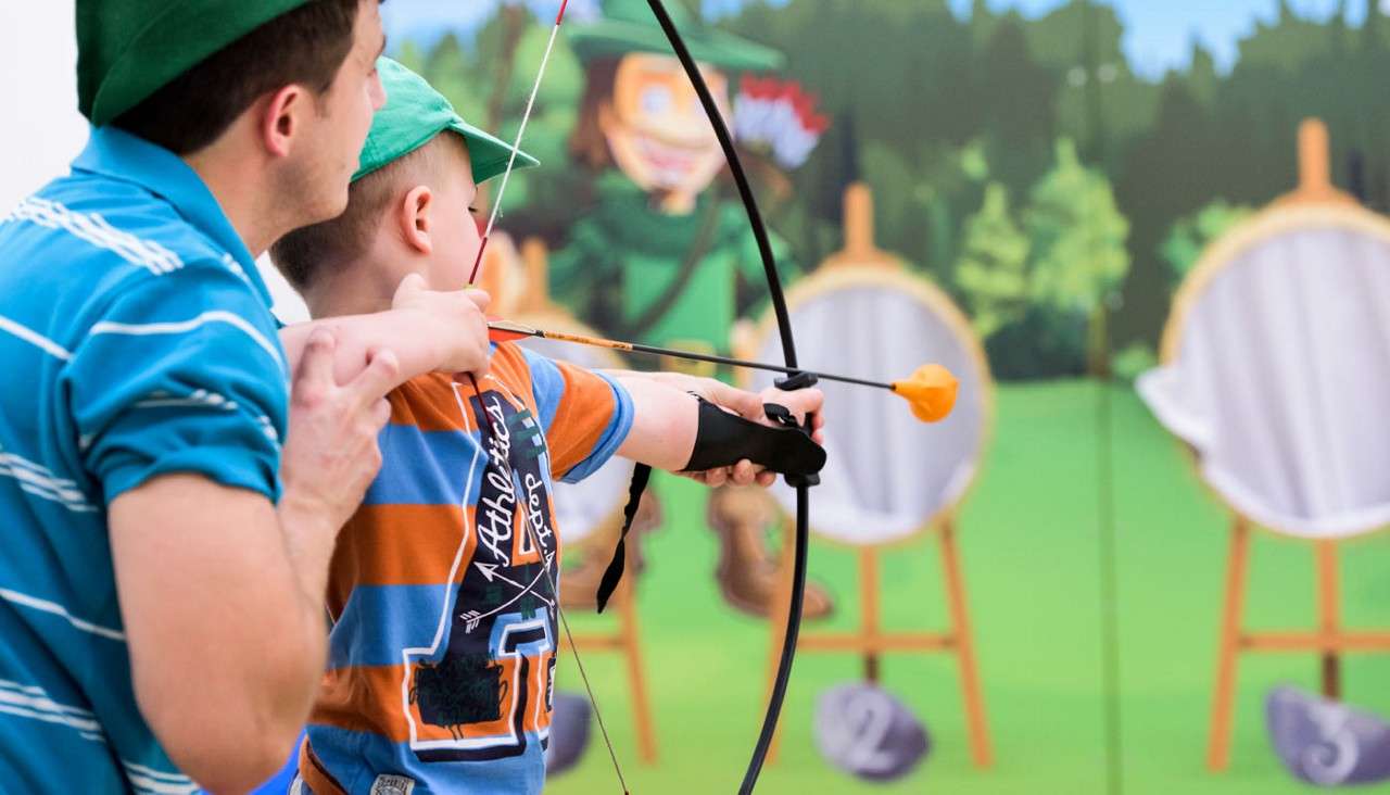 Boy archer aims a suction-cup arrow while an adult guides his stance, facing round targets on stands against a cartoon forest backdrop. Text: “Athletics,” “2,” “3.”