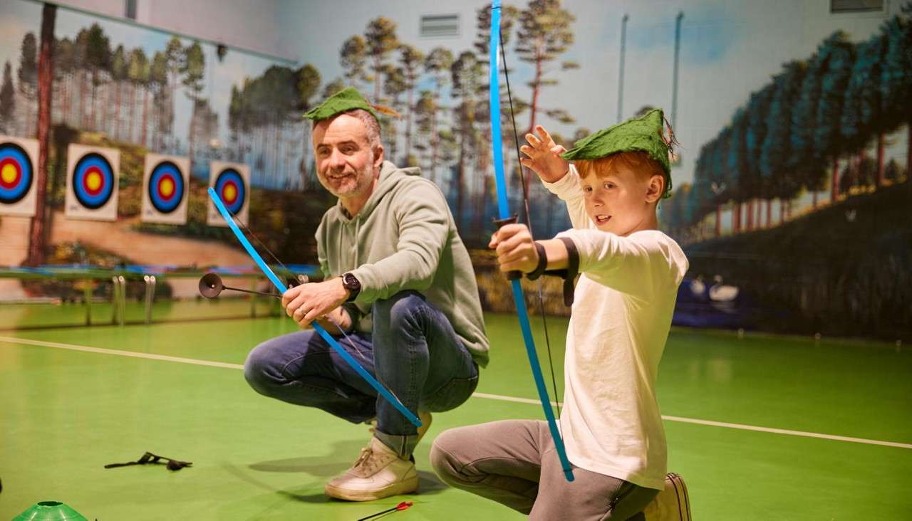 Boy archer draws a blue bow while an adult kneels beside holding another bow, coaching. Context: indoor archery range with multiple bullseye targets and forest mural background, green floor.