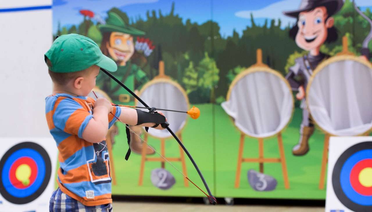 Child archer aims a toy bow with a suction-cup arrow, standing indoors before cartoon archery targets and a forest mural; numbered stones beneath targets read: 2, 3.