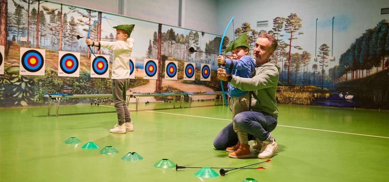Children with toy bows aim and shoot suction-cup arrows as an adult guides one. In an indoor archery range, targets line a forest-mural wall and cones mark the floor.