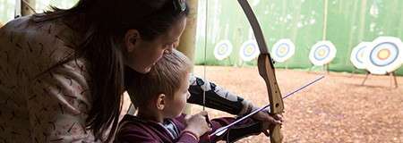 Child archer draws a bow while an adult guides their aim; in front, several circular targets stand across a leaf-covered archery range enclosed by green netting.