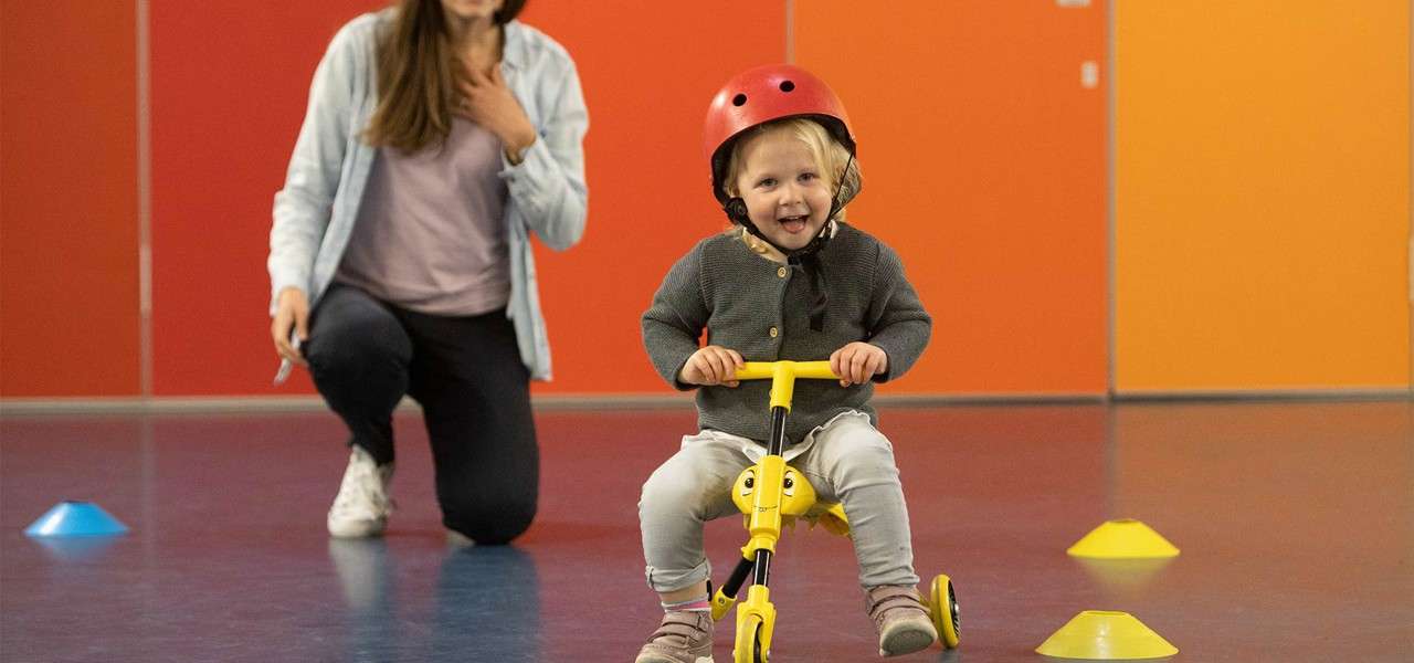 Toddler wearing a red helmet rides a small yellow tricycle, steering between plastic cones, while an adult kneels behind watching in a brightly colored indoor gym.