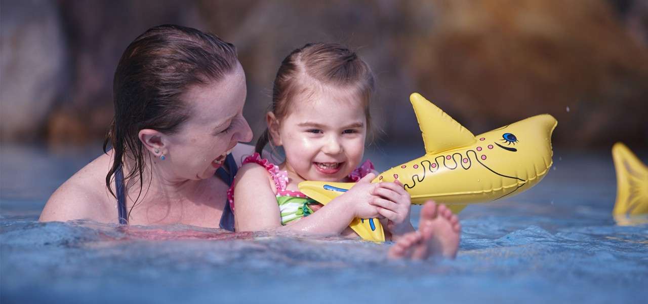 Child plays, holding a yellow inflatable shark while smiling and kicking. An adult supports them in waist-deep water, suggesting a swimming pool with a rocky, blurred background.
