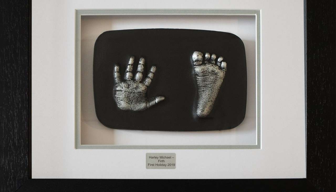 Baby handprint and footprint casts displayed side by side, mounted on a black rectangle within a white-matted black frame. Engraved plaque reads: Harley Michael – Firth; First Holiday 2019.
