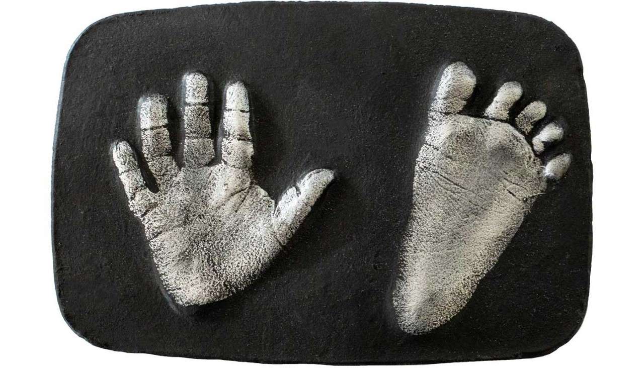Handprint and footprint pressed into a dark rectangular slab, creating pale, textured reliefs; the slab has rounded corners and smooth surrounding surface.