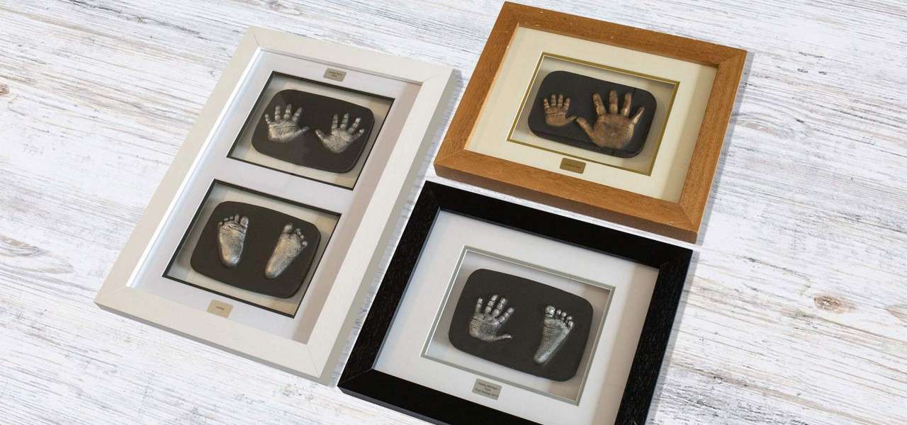 Framed casts display infant hands and feet, mounted in pairs on black backing; arranged on a light wooden surface with white, oak, and black frames. Text on small plaques is illegible.
