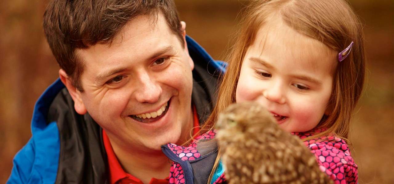 Child observes a small owl while an adult smiles nearby, both facing the bird. Context: close-up, outdoor setting with warm light and a blurred brown background.