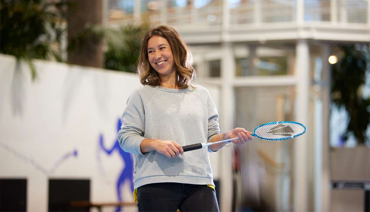 Woman holds a badminton racket and smiles, mid-gesture, as if ready to play, in a bright indoor recreation area with white walls, plants, and balcony railings blurred in background.