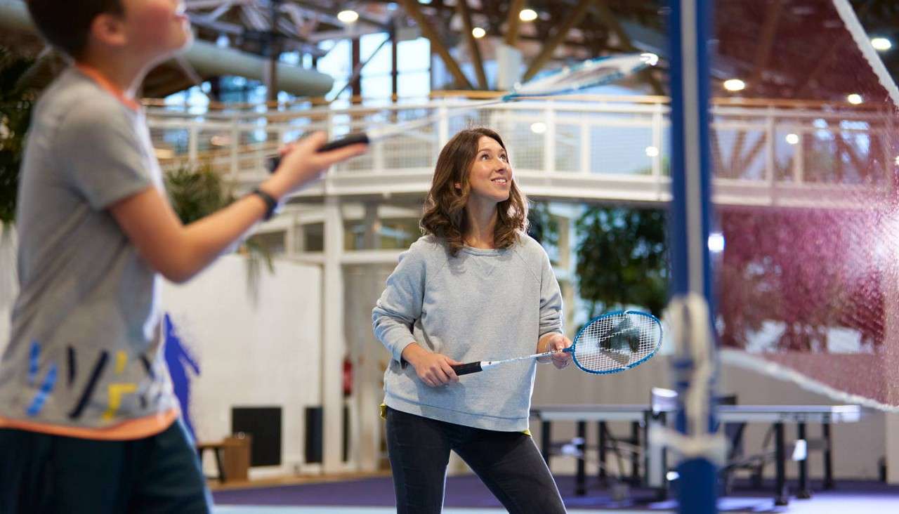 Woman with badminton racket readies to hit a shuttlecock, looking upward, while a child opponent waits nearby; indoors on a court with a net, bright lights, and other gym equipment.