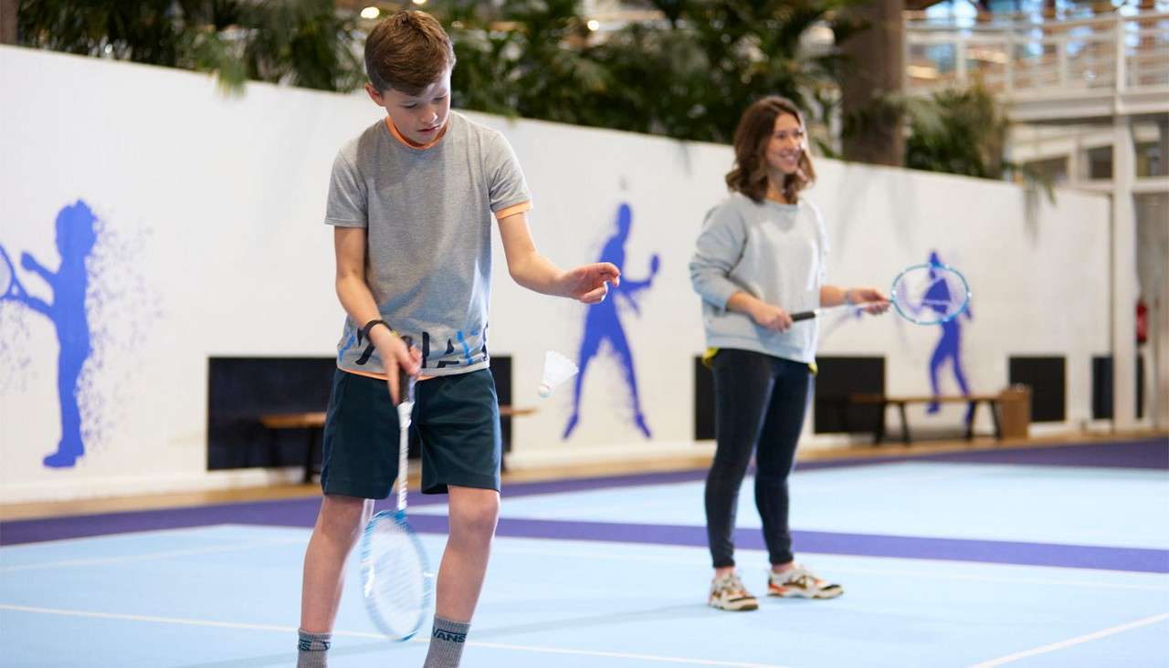 Boy practices badminton, gently tossing a shuttlecock with his racket; woman behind holds a racket, smiling. Indoor court with blue floor, wall silhouettes, benches, plants. Visible text: VANS.