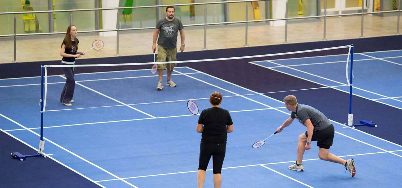 Four players rally in a doubles badminton match, swinging racquets as one lunges near the net. They play on blue indoor courts with white lines, railings and windows surrounding.