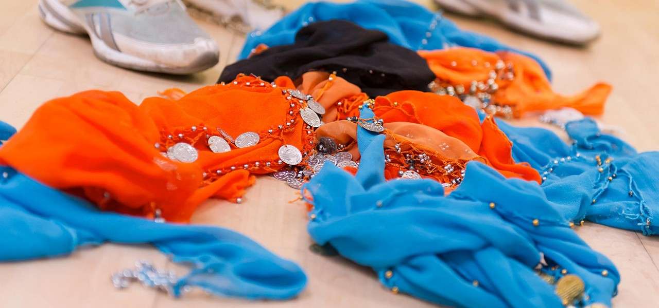 Beaded belly-dance scarves lie scattered, coins shimmering; context: a wooden studio floor, with two athletic shoes in the background.